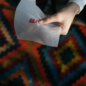 Close-up of a hand embroidering 'YES' on fabric over a colorful geometric patterned rug.