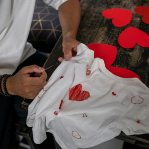 Person embroidering heart shapes on a white shirt, themed for Valentine's Day indoors.